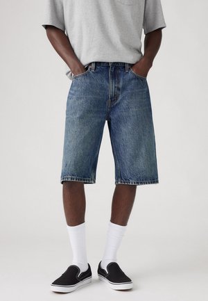 Man draagt blauw spijkerbroekshorts van gemiddelde lengte, grijs T-shirt, witte sokken en zwarte instapschoenen, staand met handen in de zakken.