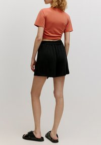 Schwarze, gefaltete Shorts mit einem elastischen Bund, kombiniert mit einem kurz geschnittenen, lachsfarbenen Baumwoll-T-Shirt und schwarzen Rutsch-Sandalen.