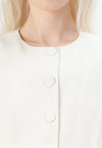 White linen garment with a rounded neckline, featuring three fabric-covered buttons down the front. Texture is subtle and natural.