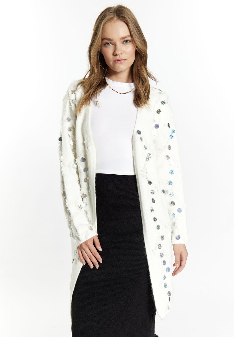 Cardigan bianco oversize con paillettes colorate e bordi sfrangiati. Presenta maniche lunghe e una vestibilità aperta. Indossato su una gonna nera aderente.