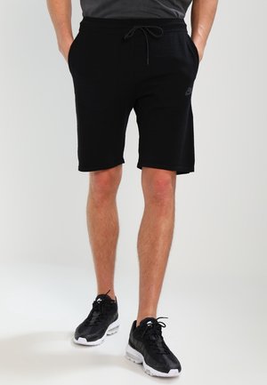 Man in zwarte trekkoordshorts, een donkergrijs shirt en zwart-witte sportschoenen, staande met handen in de zakken.