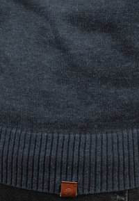 Marineblauer Strickpullover mit geripptem Saum und subtiler Textur, versehen mit einem kleinen braunen Lederschild mit rundem Logo.