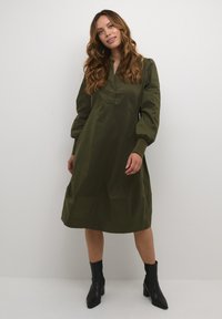 Olivovo zelené šaty s dlhými nadýchanými rukávmi, plytkým golierom a voľným strihom. Nosené s čiernymi členkovými topánkami, hladká textúra látky.