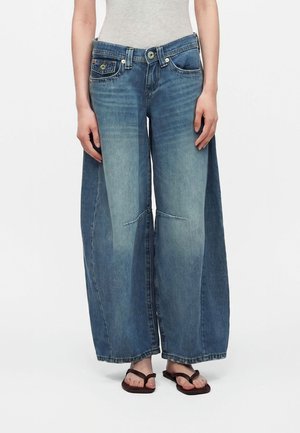 Person iført en lysegrå tanktop, brede blå jeans med falmede områder og mørke klipklappere, stående foran en ensfarvet hvid baggrund.