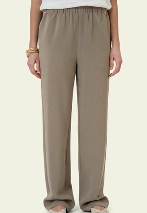 Vorderansicht von beige, locker sitzenden, hoch taillierten Hosen mit elastischem Bund, getragen mit weißem Top und Armbändern am linken Handgelenk.