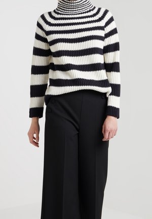 Femme portant un pull à col roulé côtelé rayé noir et blanc et un pantalon noir taille haute à jambes larges avec détails de boutons.