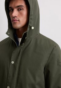Mężczyzna w oliwkowej zielonej kurtce z kapturem, zapinanej na zamek błyskawiczny i zatrzaski, widoczna część niebieskiej koszuli pod spodem, neutralne tło.
