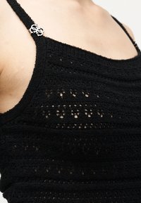 Μαύρο αμάνικο πλεκτό τοπ με λεπτές τιράντες και ασημένια διακοσμητική λεπτομέρεια με λογότυπο στην αριστερή τιράντα, φορεμένο σε γυμνό δέρμα.