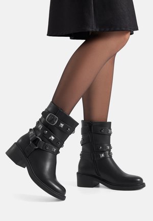 Piernas con medias negras transparentes que llevan botas de cuero negras hasta la mitad de la pantorrilla, con correas, hebillas y tachuelas de metal, sobre un fondo blanco.