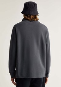 Donkergrijze langekraag top met lange mouwen, minimale branding aan de achterkant, een gladde textuur en geribbelde manchetten. Gecombineerd met een zwarte bucket hat.
