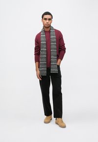 Bordowy sweter dziany z prążkowanymi mankietami, zestawiony z czarnymi spodniami o prostych nogawkach; szary szal w paski; beżowe trampki. Minimalistyczny design.