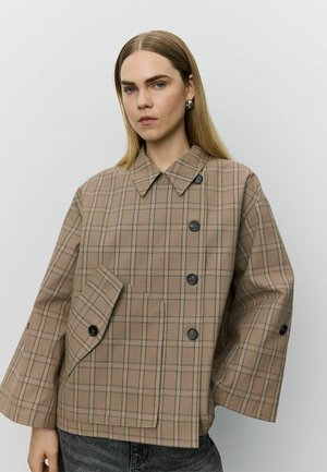 Femme aux cheveux blonds et raides portant une veste oversize à carreaux beige avec de gros boutons et un large col sur un fond uni.