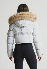 Balta īsā pūkainā jaka ar bēšas mākslīgās kažokādas kapuci. Iespējas ar kviltēto dizainu un elastīgām piedurknēm. Valkāta virs melnām legingām.