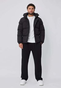 Doudoune noire avec des boutons pression, une capuche et un design matelassé, portée sur un t-shirt blanc et associée à un pantalon noir ample et des baskets blanches.