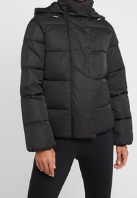 Giacca a blocchi nera con cappuccio, caratterizzata da una texture trapuntata, un taglio dritto e dettagli in metallo argentato sul colletto e sulla cerniera.