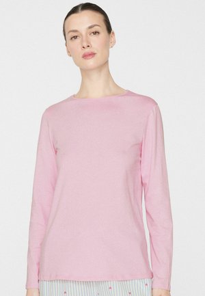 Femme aux cheveux foncés attachés, portant une chemise rose pâle à manches longues et un pantalon rayé avec de petits motifs en forme de cœur, debout devant un fond blanc.