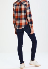 Camisa de franela a cuadros en óxido, azul marino y crema; combinada con vaqueros ajustados oscuros y zapatillas blancas. Presenta puños con botones y dobladillo curvado.