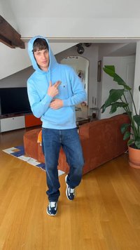 Mladý muž v modré mikině s kapucí a džínách ukazuje na hruď, stojí na dřevěné podlaze v obývacím pokoji s oranžovou pohovkou a velkou květináčem s rostlinou.