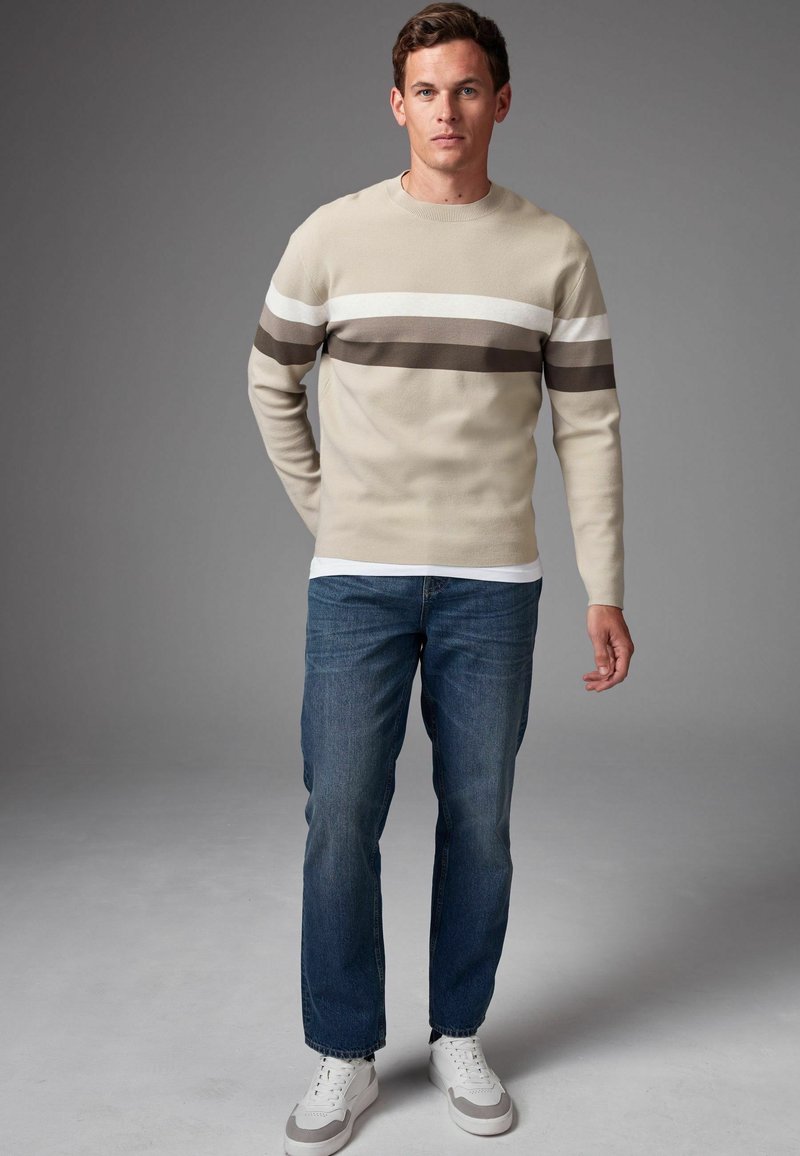 Beige Pullover mit Rundhalsausschnitt und braunen horizontalen Streifen, kombiniert mit blauen Jeans und weißen Sneakers, verfügt über eine glatte Textur und eine lässige Passform.