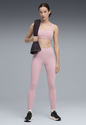 Donna con reggiseno sportivo rosa e leggings che tiene un asciugamano scuro sulle spalle, in piedi su uno sfondo grigio sfumato e indossa sneakers nere.