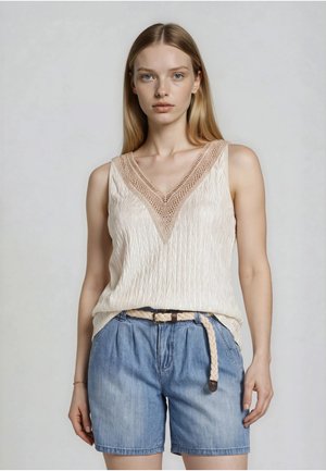 Junge Frau trägt eine cremefarbene, strukturierte ärmellose Bluse mit gehäkeltem V-Ausschnitt und hellblaue Denim-Shorts mit geflochtenem Gürtel, steht vor einem schlichten Hintergrund.