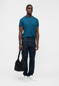 Mann som står iført t-skjorte i teal, mørke jeans og hvite joggesko, holder en svart skulderveske mot en ensfarget hvit bakgrunn.