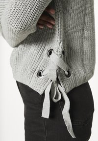 Jasnoszary sweter dzianinowy o luźnym kroju, z bocznym wiązaniem na sznurowadło i oczkami oraz miękką fakturą. Połączony z ciemnymi czarnymi spodniami.