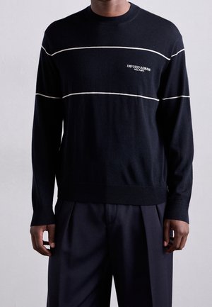 Man draagt een zwarte trui met lange mouwen met witte horizontale strepen en het "Emporio Armani Milano" logo op de borst, gecombineerd met een donkere broek.