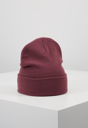 Tummanpunainen neulottu pipo, jossa on käännetty reunus, asetettuna valkoisen jalustan päälle vaaleanharmaata taustaa vasten.