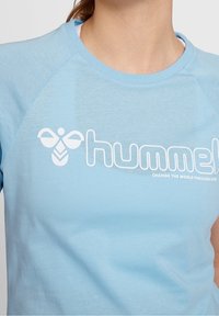 Jasnoniebieska koszulka z krótkim rękawem, okrągłym dekoltem i rękawami raglanowymi. Z przodu znajduje się białe logo i tekst. Miękka struktura tkaniny.