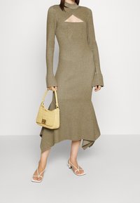 Femme portant une robe midi côtelée vert olive à manches évasées et ourlet asymétrique, tenant un sac beige et portant des talons blancs à lanières.