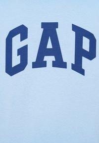 T-shirt en coton bleu clair avec de grandes lettres bleu marine formant "GAP" sur la poitrine. Texture douce et design casual.
