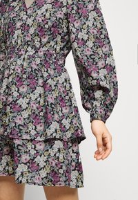 Kvinna iklädd en långärmad blommig klänning med samlad midja och lager-på-lager kjol i lila, vitt, grönt och blått på svart tyg.