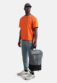 Grå og sort rygsæk med et trekantet design, lynlås og logo. Model i orange skjorte, grå bukser og lyse sneakers.