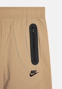 Calções bege com uma cintura elástica, apresentando um bolso com fecho éclair preto na frente e um logotipo preto da Nike na parte inferior. Textura suave.