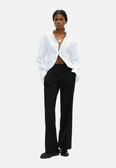 Witte oversized shirt met open voorkant, zwarte tailored broek en zwarte schoenen. Gestructureerde stof met een hartvormige hangerketting.