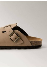 Sandal din suede bej, cu formă curbată, curea ajustabilă cu o cataramă din bronz și talpă din plută, având o talpă texturată din cauciuc negru.
