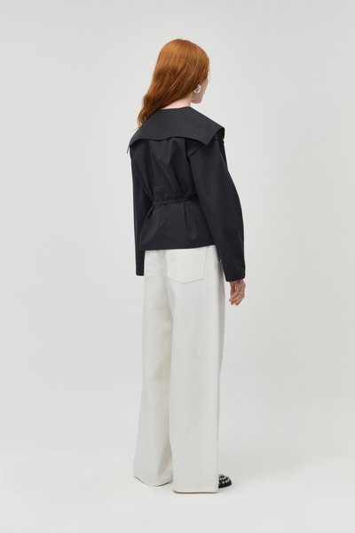 Femme aux cheveux roux portant un haut noir à manches longues avec un large col et un pantalon blanc à jambes larges, debout de dos sur un fond uni.