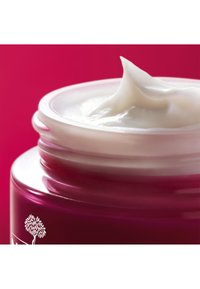 Różowy szklany słoik z białą kremową teksturą wystającą od góry. Słoik ma gładkie wykończenie i kwiatowy detal logo po boku.