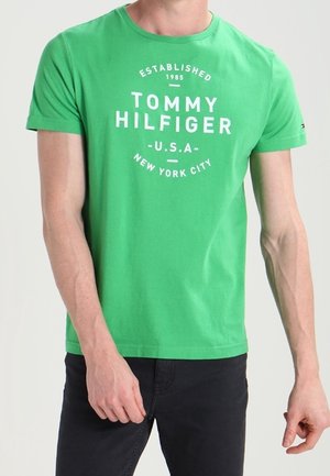 Homme portant un t-shirt vert Tommy Hilfiger avec le texte blanc « Established 1985 Tommy Hilfiger U.S.A New York City » et un pantalon noir.