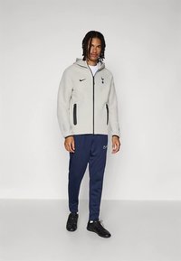 Gaiši pelēks fliša žakete ar melnu rāvējslēdzēju un kabatām, tumši zili sporta bikses, melni sporta apavi ar logotipa akcentiem.