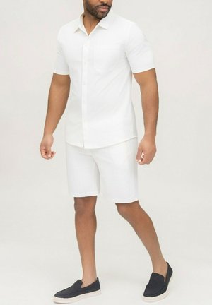Homme portant une chemise blanche à manches courtes avec boutons, un short blanc jusqu'aux genoux et des chaussures noires sans lacets, sur un fond uni.