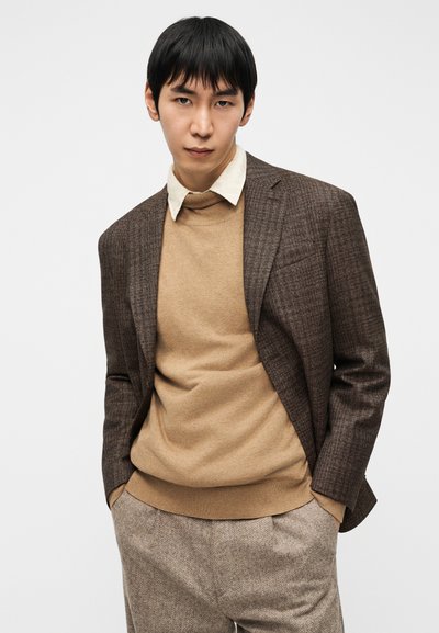 Καφέ καρό σακάκι πάνω από μπεζ πουλόβερ με λαιμόκοψη, συνδυασμένο με μπεζ παντελόνι με σχέδια. Ορατός λευκός γιακάς. Λείες υφές και ραμμένη εφαρμογή.