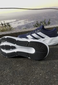 Navy blue athletic shoes with white stripes, one showing textured black and white sole, placed on asphalt near a stone edge with blurred landscape background.