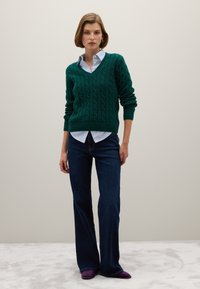 Grüner Kabelstrickpullover mit V-Ausschnitt, über einem hellblau-weiß gestreiften Hemd getragen. Kombiniert mit dunkelblauen Schlagjeans und lila Schuhen.