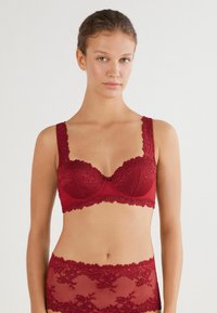 Czerwony koronkowy biustonosz z fiszbinami i wyprofilowanymi miseczkami, z falbankowymi brzegami i dekoracyjnymi akcentami. Miękka tekstura i skomplikowane kwiatowe wzory.