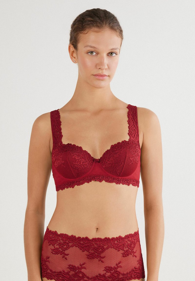 Czerwony koronkowy biustonosz z fiszbinami i wyprofilowanymi miseczkami, z falbankowymi brzegami i dekoracyjnymi akcentami. Miękka tekstura i skomplikowane kwiatowe wzory.