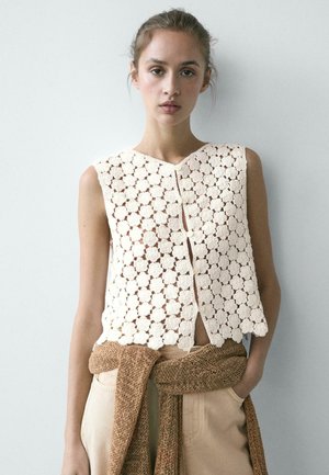 Giovane donna che indossa un top senza maniche bianco con motivo floreale all'uncinetto e pantaloni beige, con un maglione marrone a maglia legato in vita.