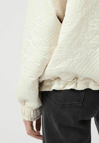 Giacca trapuntata bianca sporca con polsini a coste, caratterizzata da delicati motivi floreali e un orlo arr gathered, abbinata a jeans grigio scuro.
