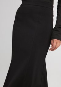 Schwarzer langer Rock aus einem glatten Stoff, mit einer taillierten Taille und einem ausgestellten Design. Seitenschlitz und sichtbare Nahtakzente.